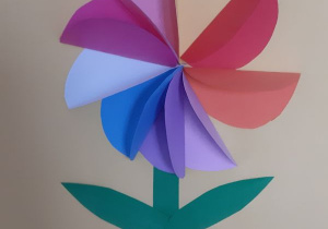 kolorowy kwiatek z kolorowego papieru wykonany technika origamii, z dwoma listkami i łodygą na kremowej kartce